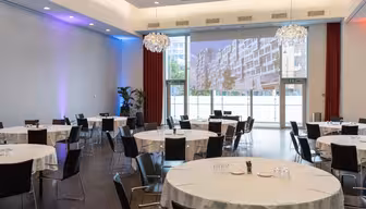 A modern conference room with round tables covered in white tablecloths and surrounded by black chairs. Glasses and plates are set on the tables. Large windows and a projected cityscape image enhance the bright and spacious atmosphere.