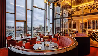 A modern, luxurious restaurant interior with large windows showcasing a cityscape. The space is adorned with red chairs, round tables set for dining, wine racks, and warm lighting from elegant pendant lights.