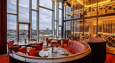 A modern, luxurious restaurant interior with large windows showcasing a cityscape. The space is adorned with red chairs, round tables set for dining, wine racks, and warm lighting from elegant pendant lights.
