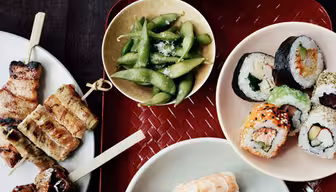 A variety of Japanese dishes are displayed. Sushi rolls, nigiri with shrimp and tuna, and skewered grilled meats are on white plates. A red tray holds edamame in a bowl and a bowl of rice. Chopsticks rest on the tray.
