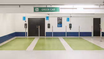 A parking garage featuring multiple green electric vehicle charging spots with a prominent Green Car sign above. Black chargers are mounted on the wall, and the area is marked by bright green paint on the floor.